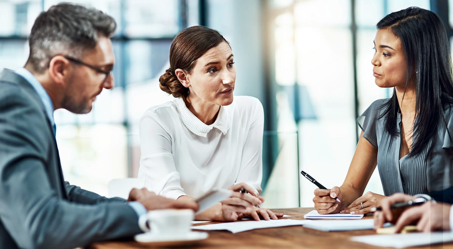 trois professionnels en tenue de travail engagés dans une discussion animée autour d'une table de conférence en bois dans un bureau moderne.
